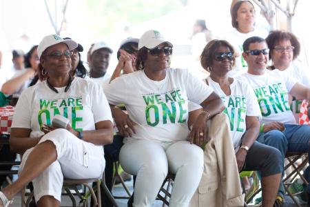 A group of people is seated under a tent, all wearing matching white t-shirts with green text that reads "We Are One" and caps with the Chicago Housing Authority logo. The group includes both men and women, who appear to be engaged in an event or presentation. They are smiling and relaxed, with some wearing sunglasses. The background shows more people in similar attire, creating a sense of unity and community.